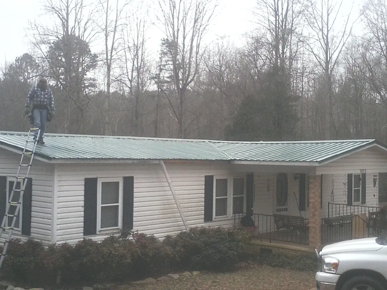 Worker on a ladder beside a new green metal roof on a ranch home in Camden